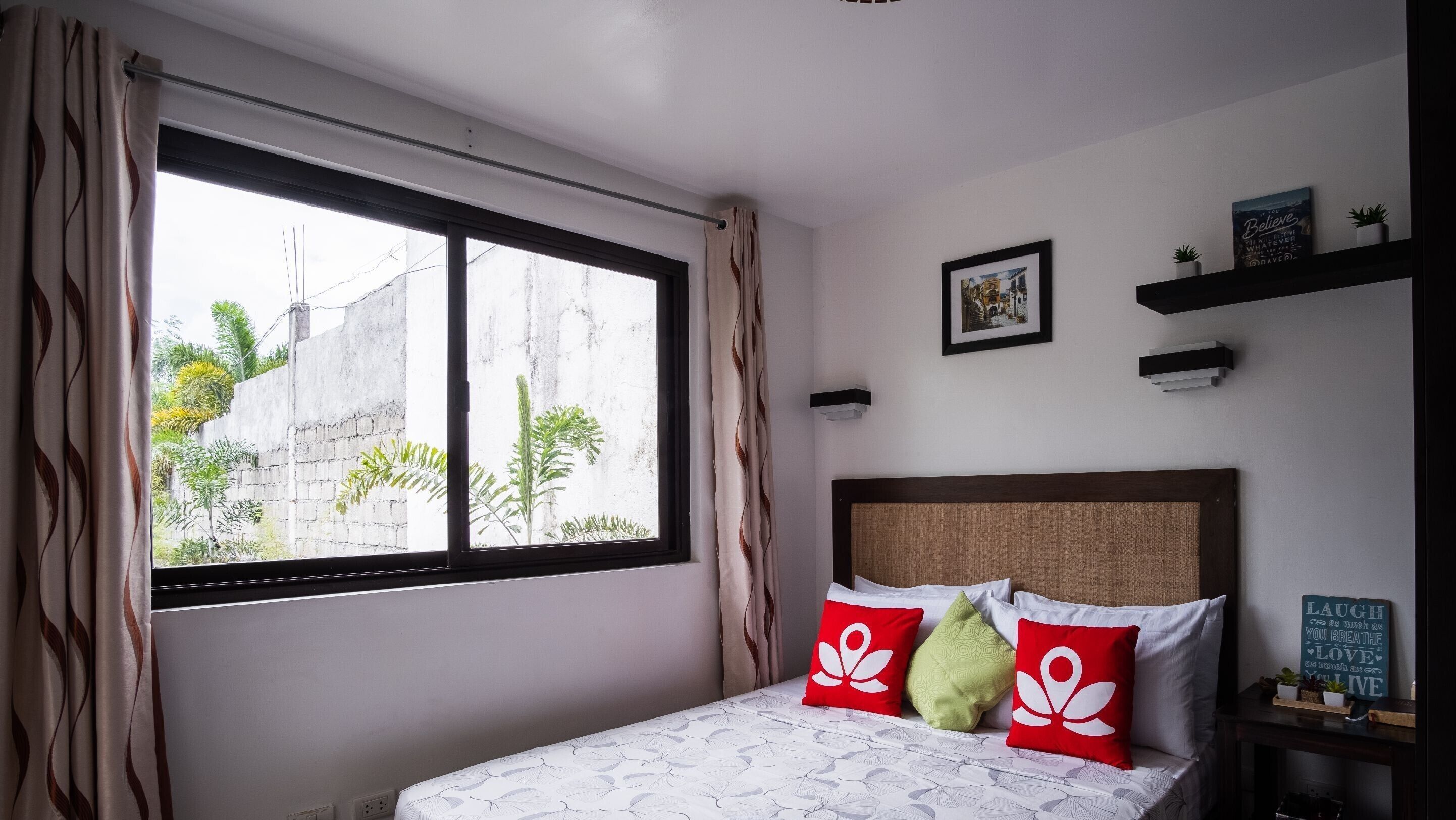 A bedroom with a bed, a window with curtains, a bedside table with a plant, and a wall with a framed picture and a sign.
