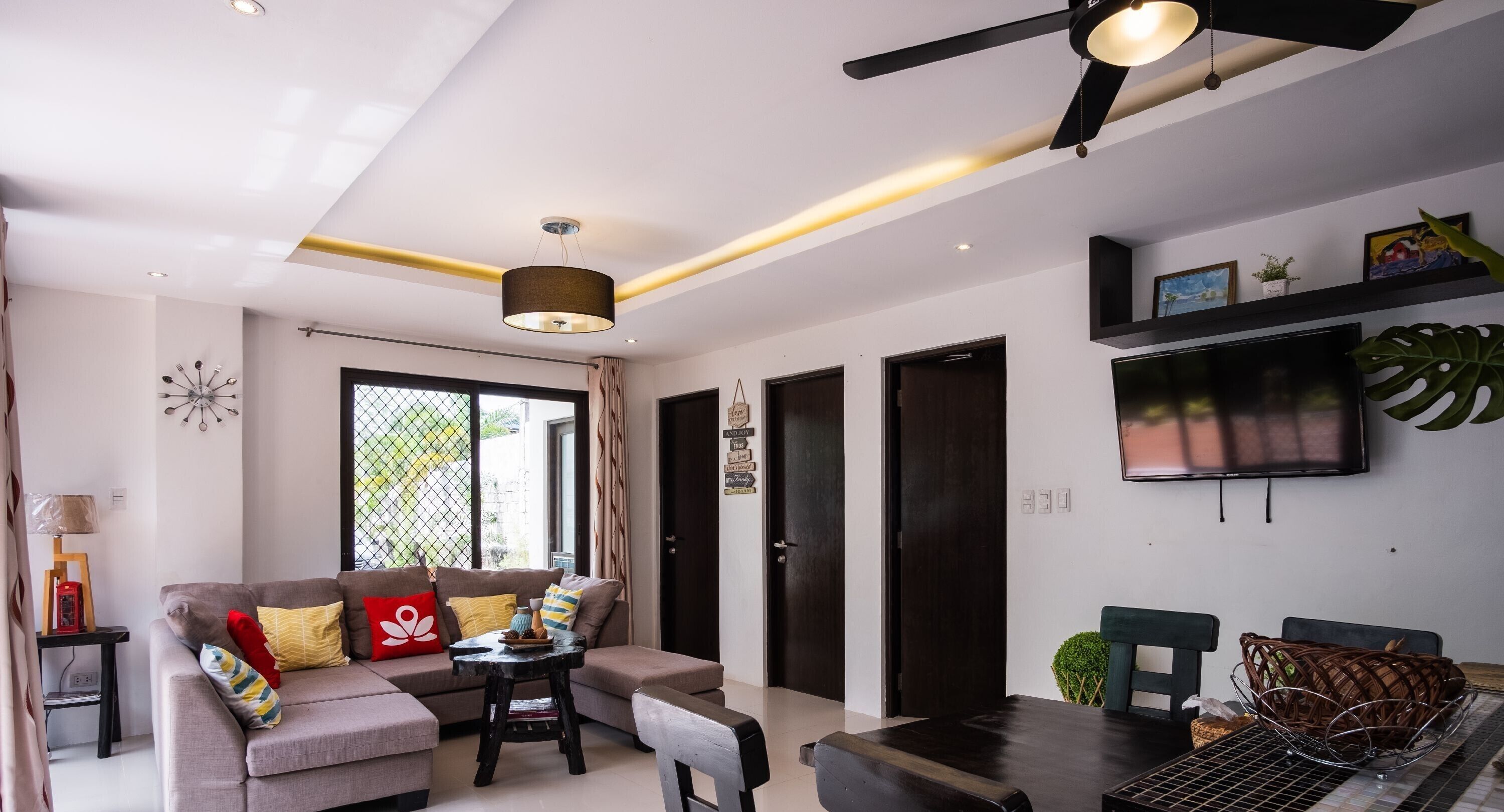 A modern living room with a sofa, coffee table, and dining table. There is a ceiling fan, a TV mounted on the wall, and a window with curtains.