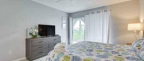 Une chambre à coucher avec un lit, un téléviseur posé sur une commode, un ventilateur de plafond et une fenêtre avec des rideaux.