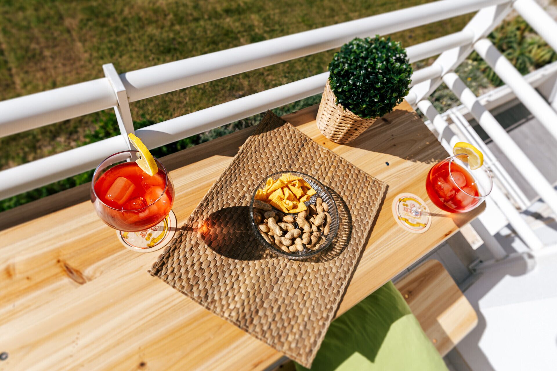 A wooden deck with a woven placemat, two drinks with lemon slices, a bowl of peanuts, and a small potted plant.