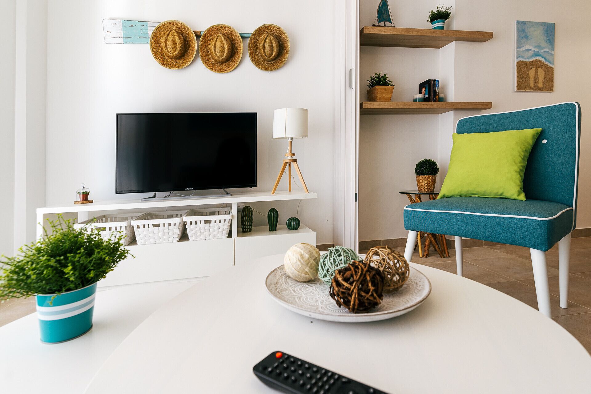 Una sala moderna con televisor de pantalla plana, un sofá azul con un cojín verde, una mesa de centro blanca con objetos decorativos y un mueble de pared para el televisor.