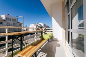 A balcony with a wooden bench, a small table with a basket and a glass, and a view of a white building with balconies. - Baleal Beachfront Apartment (Ferrel)