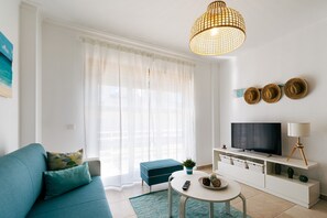 A modern living room with a teal sofa, a round coffee table, a TV on a white stand, and a large window with sheer curtains. - Baleal Beachfront Apartment (Ferrel)