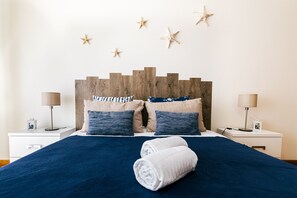 A neatly made bed with a dark blue bedspread, two pillows, and a rolled white towel. There are starfish decorations on the wall and two bedside lamps on either side of the bed.