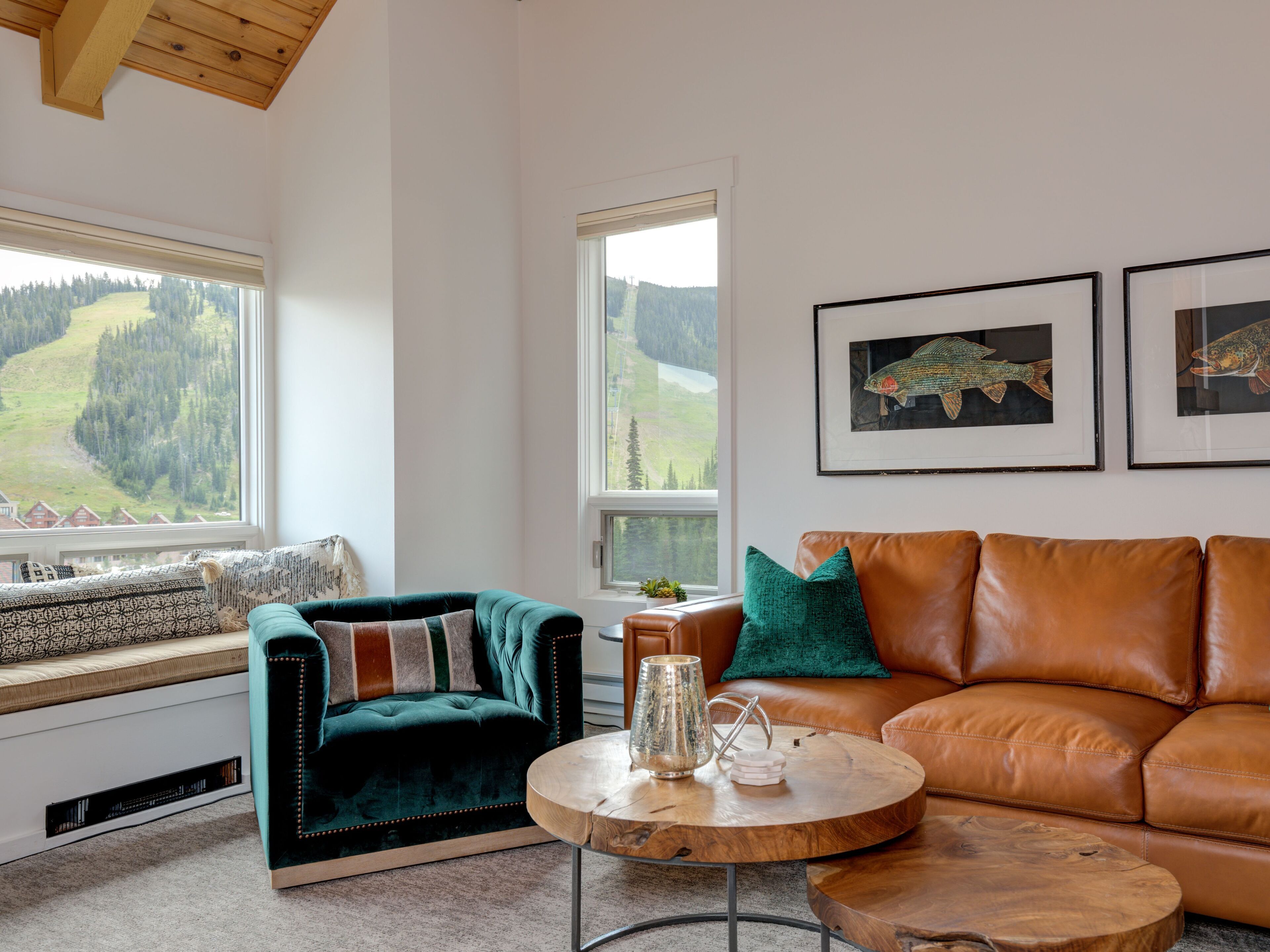 Un salon chaleureux avec un canapé en velours vert, un divan en cuir marron et une table basse ronde en bois. Deux fenêtres offrent une vue sur un paysage boisé.