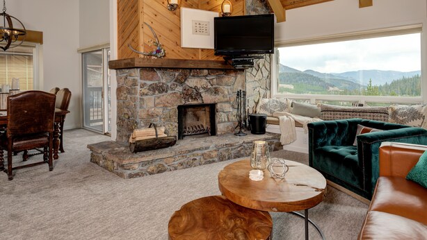 Una sala de estar acogedora con chimenea de piedra, vigas de madera en el techo, un televisor y vistas a las montañas a través de un gran ventanal.