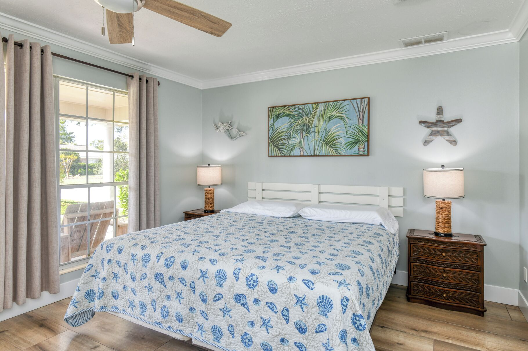 Un dormitorio con cama, dos mesitas de noche, ventilador de techo, ventana con cortinas y decoración en la pared.