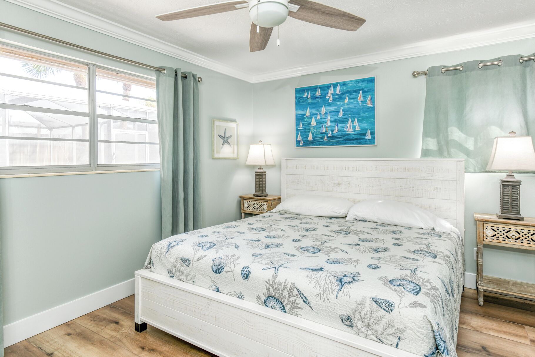 A bedroom with a bed, a ceiling fan, a window with curtains, a painting, and a nightstand with a lamp.