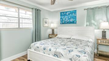 A bedroom with a bed, a ceiling fan, a window with curtains, a painting, and a nightstand with a lamp.