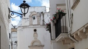 Una calle estrecha con adoquines, edificios blancos, una iglesia con campanario y macetas colgantes de flores.