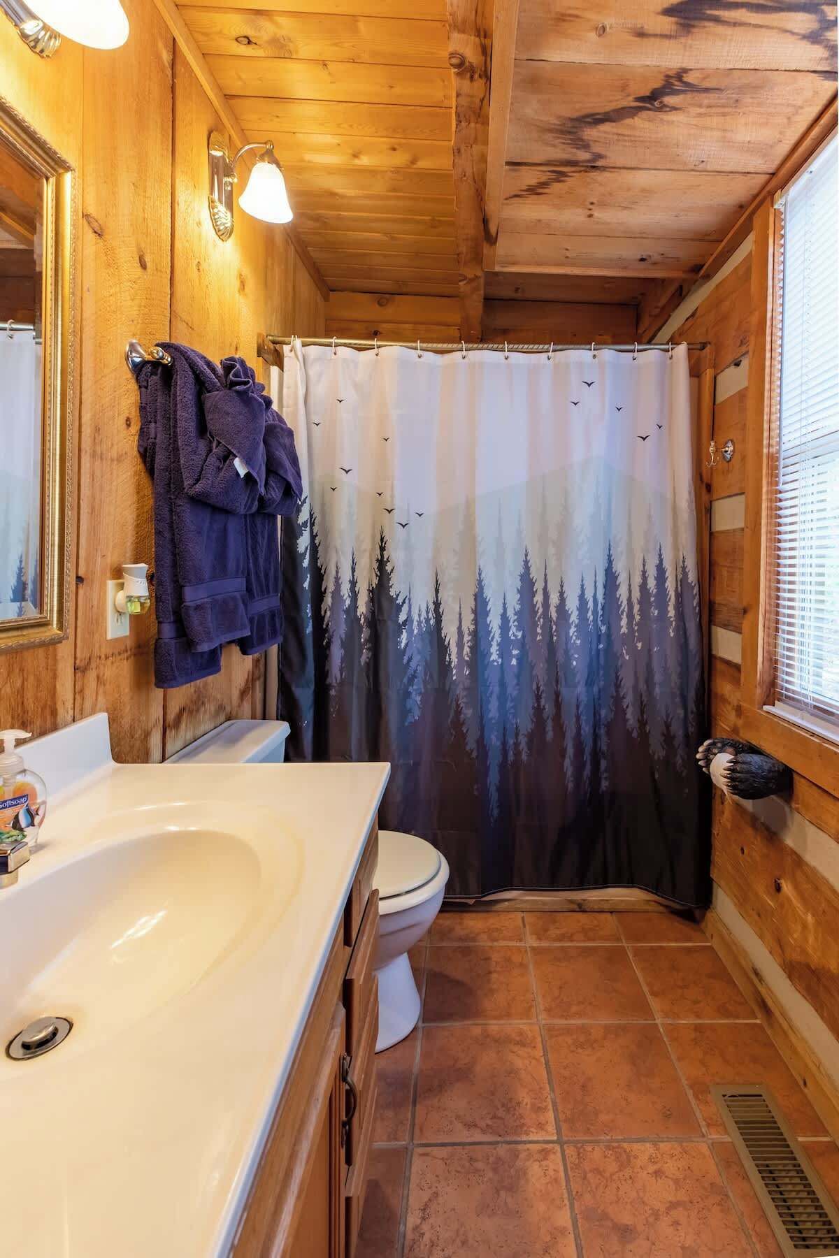 Une salle de bain rustique, avec des murs en bois, un lavabo blanc, des toilettes et un rideau de douche orné d’un motif de forêt.