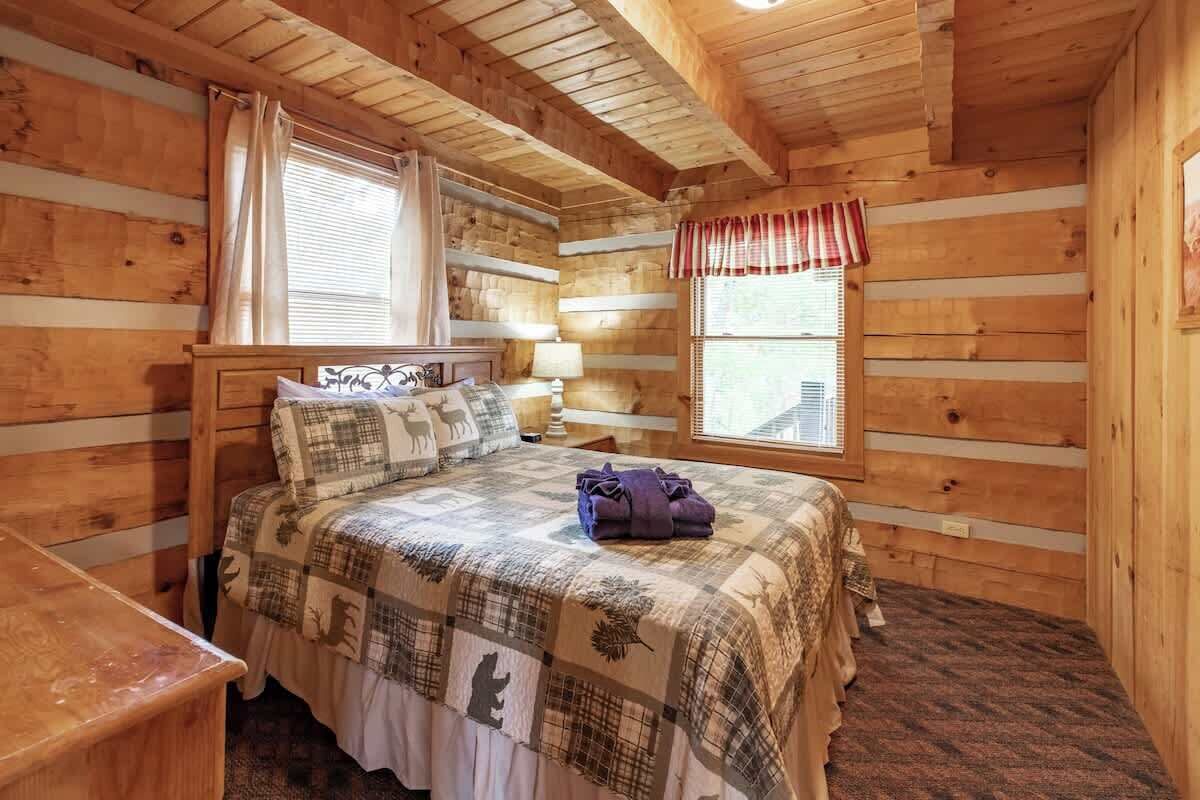 Ein Zimmer in einer Holzhütte mit einem Bett, zwei Fenstern, einem Nachttisch und einem roten Vorhang.