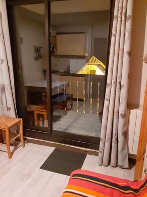 A room with a sliding glass door leading to a balcony, a small wooden stool, a colorful striped blanket, and a patterned curtain. - Homerez - Studio in Allos - mountain view (Allos)