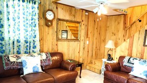 A wooden-paneled room with two brown leather sofas, a wooden staircase, a clock, and a ceiling fan.