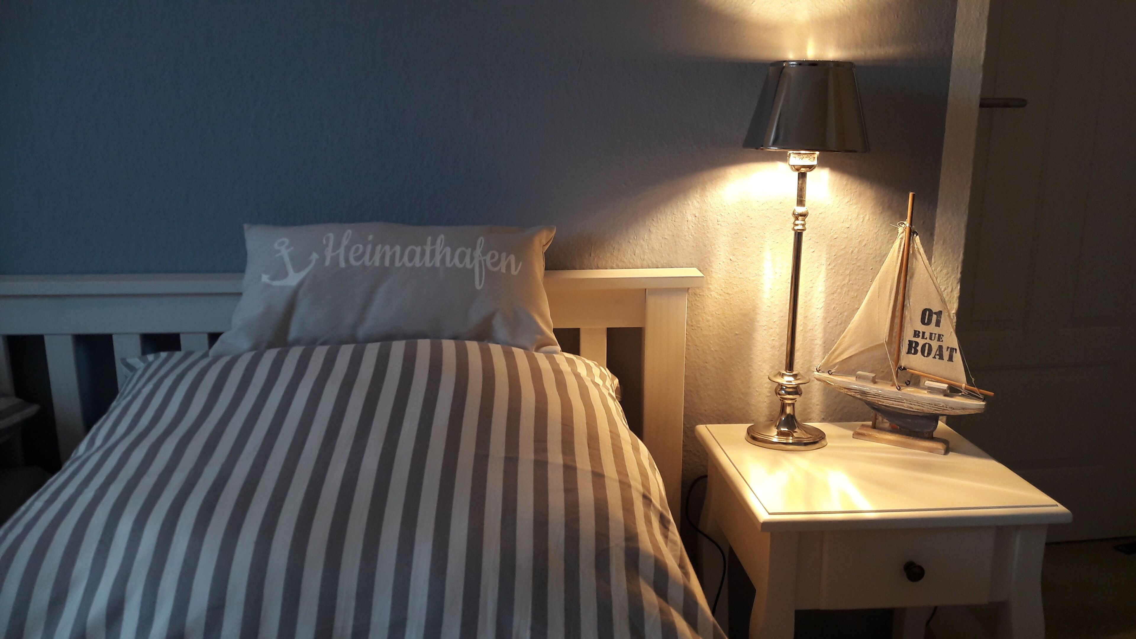 Ein Bett mit gestreifter Bettwäsche, ein Kissen mit dem Schriftzug "Heimathafen", eine Lampe und ein kleines Modell eines Segelboots auf einem Nachttisch.
