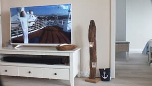 A television on a white entertainment unit with a wooden bowl on top, a candle holder with a lit candle, and a small bag with an anchor design. - Sea view, balcony, 3 room apartment (Büsum)