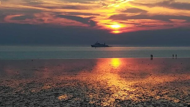 A sunset over a calm sea with a ship in the distance and a beach in the foreground. - Sea view, balcony, 3 room apartment (Büsum)