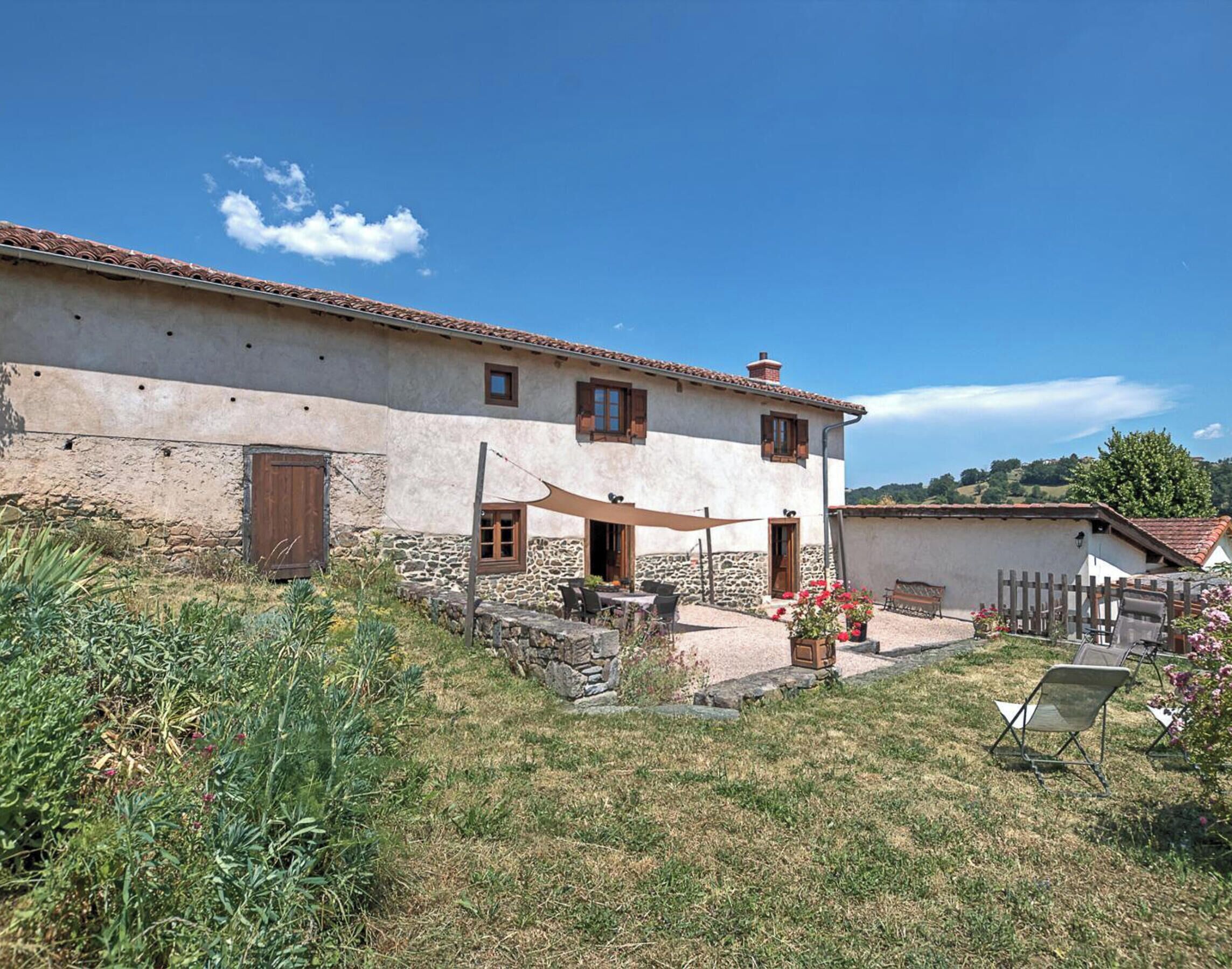 Un bâtiment à deux étages avec une façade en pierre, une porte en bois et un patio aménagé avec du mobilier d’extérieur.