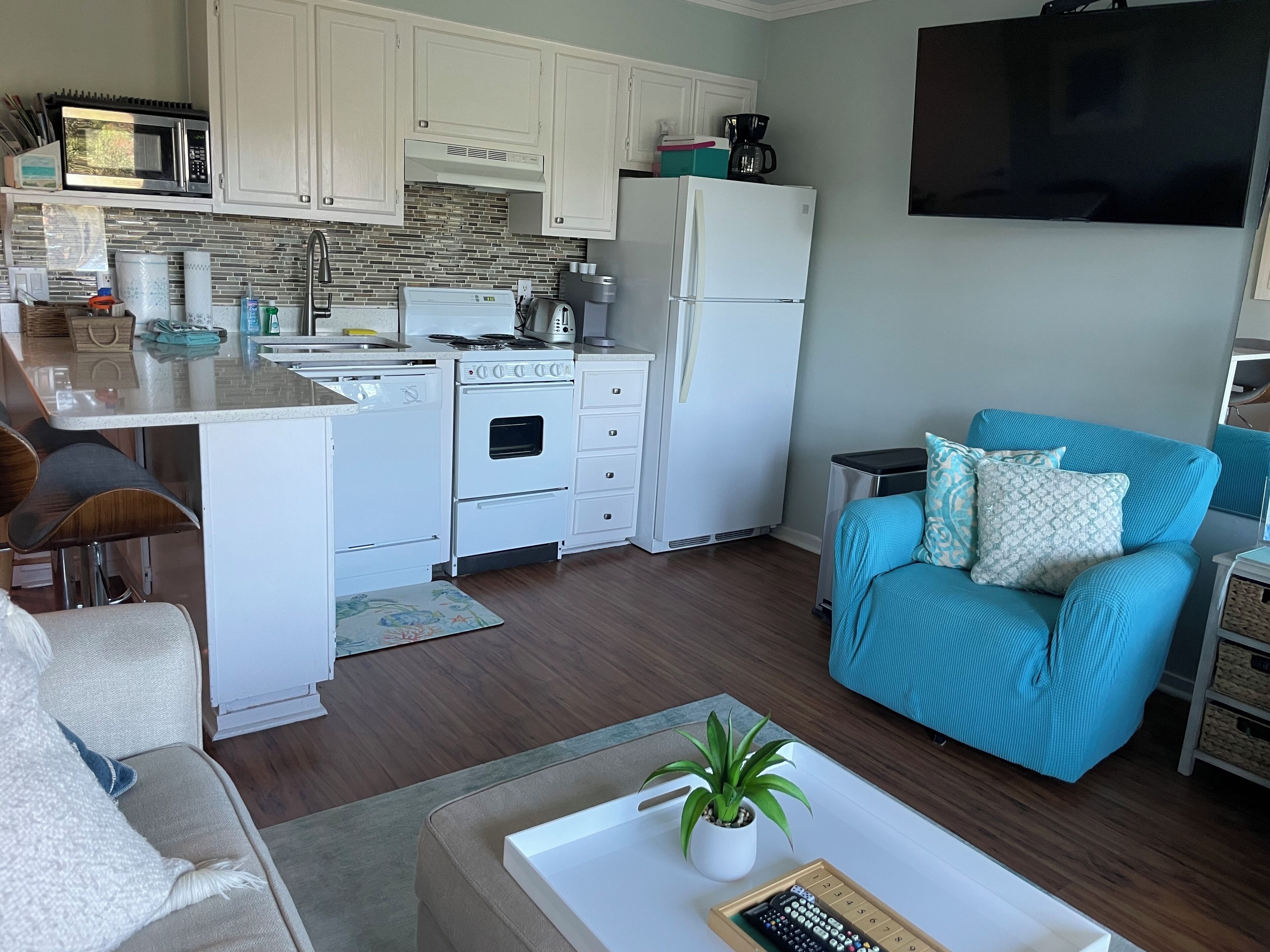 Een compacte keuken met witte kastjes, een magnetron, een fornuis en een koelkast. Er is een klein eiland met een spoelbak en een koffiezetapparaat. In de woonkamer staan een teal kleurige bank en een witte fauteuil, met een kleine tafel en een plant.
