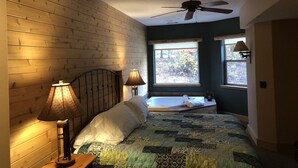 A bedroom with wooden walls, a bed with a quilt, two bedside lamps, a ceiling fan, and a window with a view of trees. - Year round family fun (New Era)
