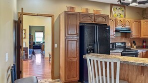 Une cuisine avec des meubles en bois, un réfrigérateur noir, une cuisinière et un coin bar avec un tabouret blanc.