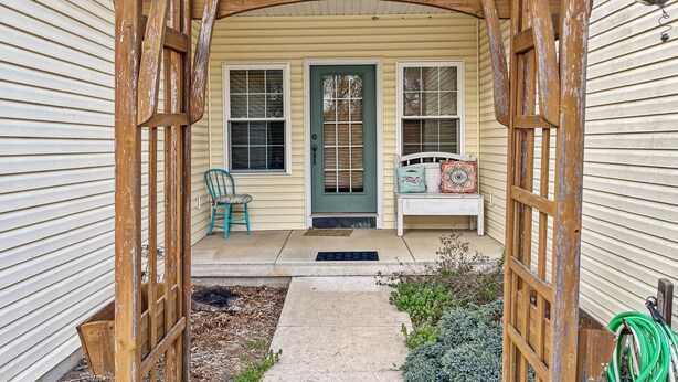 Une pergola en bois marque l’entrée d’une maison dont la porte est peinte en vert. On y trouve également un banc et une chaise à l’extérieur.