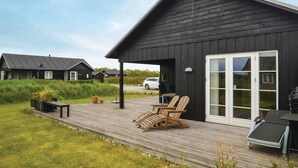 En sort bygning med træterrasse, udendørs siddepladser og en hvid bil parkeret i nærheden.