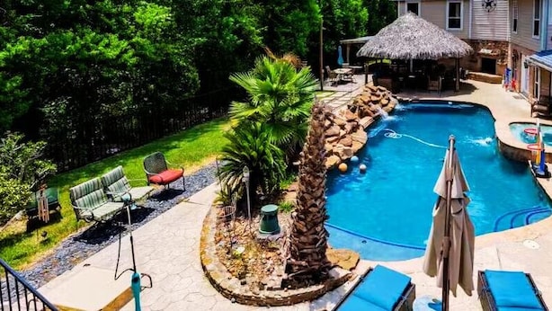 A backyard with a swimming pool, lounge chairs, a hot tub, and a thatched-roof structure.
