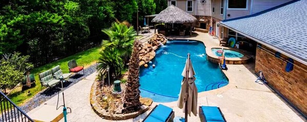 Un patio trasero con piscina, sillas de descanso, jacuzzi y una estructura de techo de paja.