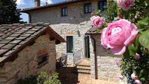 A stone building with a tiled roof, a small porch with a table and chairs, and a garden with pink roses. - Spacious home on beautiful Tuscan vineyard.  Near Florence. (Pelago)