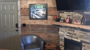 A cozy living room with a stone fireplace, a mounted flat-screen TV, a framed picture on the wall, and a leather armchair. - #4 Fall Creek Cabins family friendly 1 mile from Lake Norfork , Mtn. Home Ar. (Mountain Home)