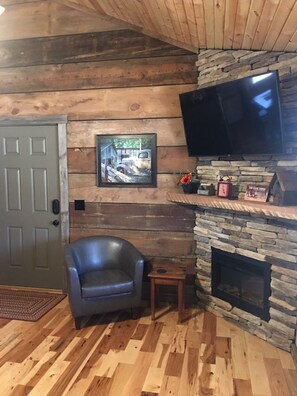 A cozy living room with a stone fireplace, a mounted flat-screen TV, a framed picture on the wall, and a leather armchair. - #4 Fall Creek Cabins family friendly 1 mile from Lake Norfork , Mtn. Home Ar. (Mountain Home)