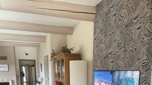 Un salón con suelo de baldosas, un mueble de madera, una estantería, un sofá con un cojín amarillo y un televisor de pared que muestra una vista urbana.