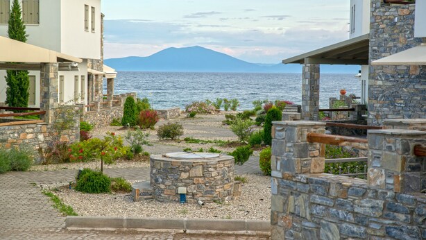 Una propietat costanera d'arquitectura de pedra, un jardí amb plantes variades i una vista al mar i una muntanya de fons.