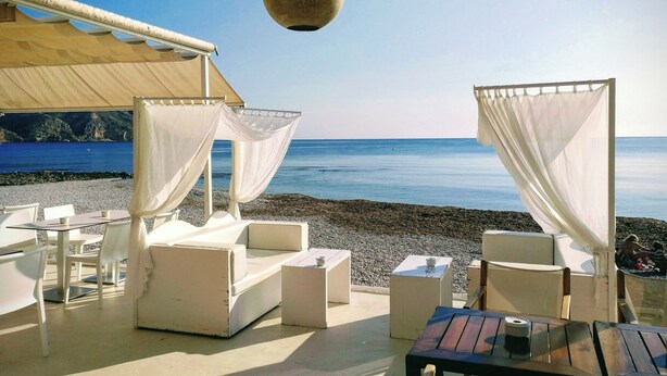 A beachside seating area with white cushioned chairs and tables, a canopy, and a hanging light fixture.