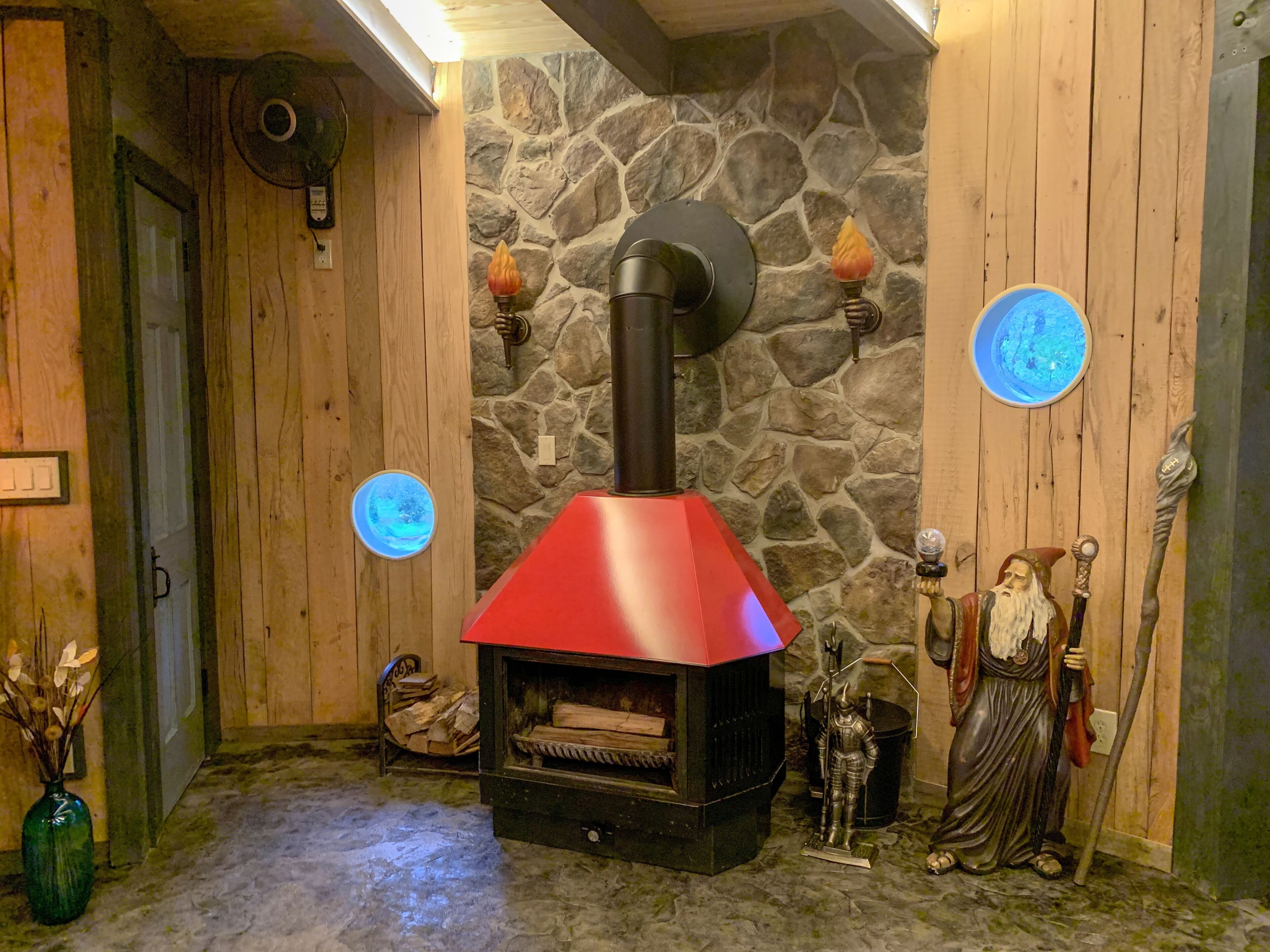 Una stanza rustica con una parete in pietra, rivestimenti in legno, un camino a legna rosso, una figura decorativa e un lampadario blu a forma circolare.