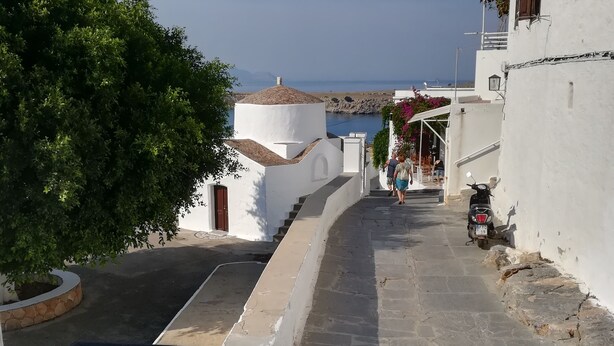 A narrow street with white buildings, a clear view of the sea, and a balcony with a table and chairs.
