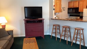 A compact living space with a television on a wooden cabinet, a sofa, a coffee table, and a kitchen area with stools. - 1BR condo near Recreation Center, pool and hot tub (North Woodstock)