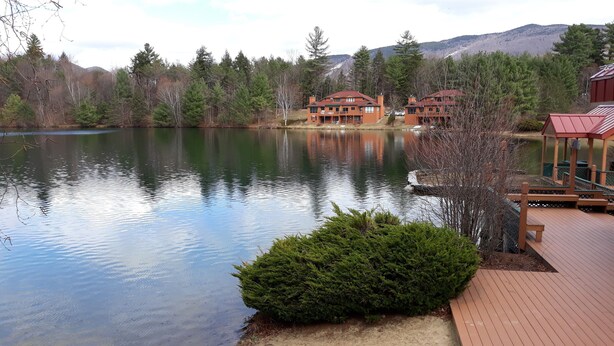 A lakeside property with a wooden deck, a small pond, and a backdrop of trees and mountains. - 1BR condo near Recreation Center, pool and hot tub (North Woodstock)