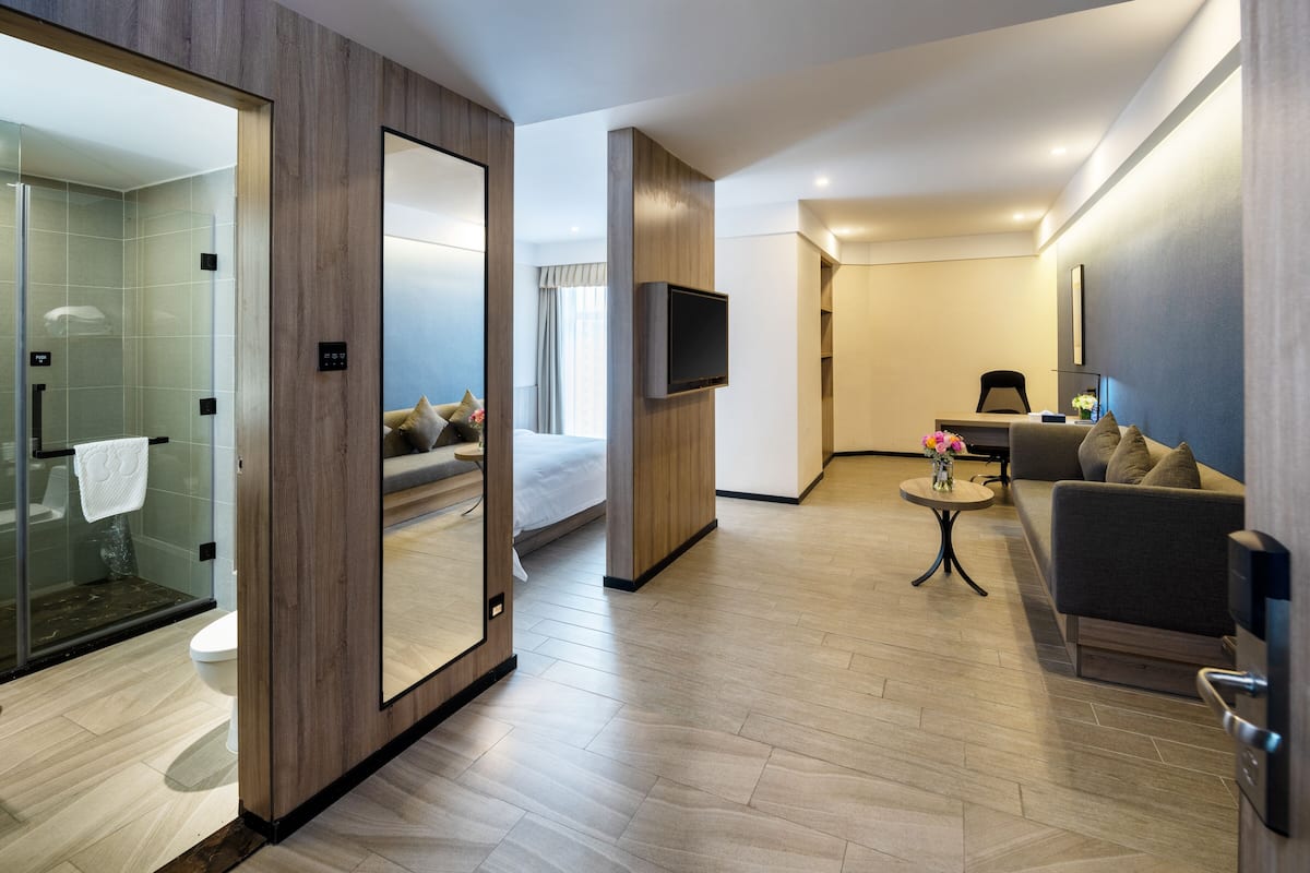 Una habitación de hotel moderna con cabina de ducha de vidrio, cama con cabecero, zona de estar con sofá y una mesita, y un televisor de pantalla plana montado en la pared.
