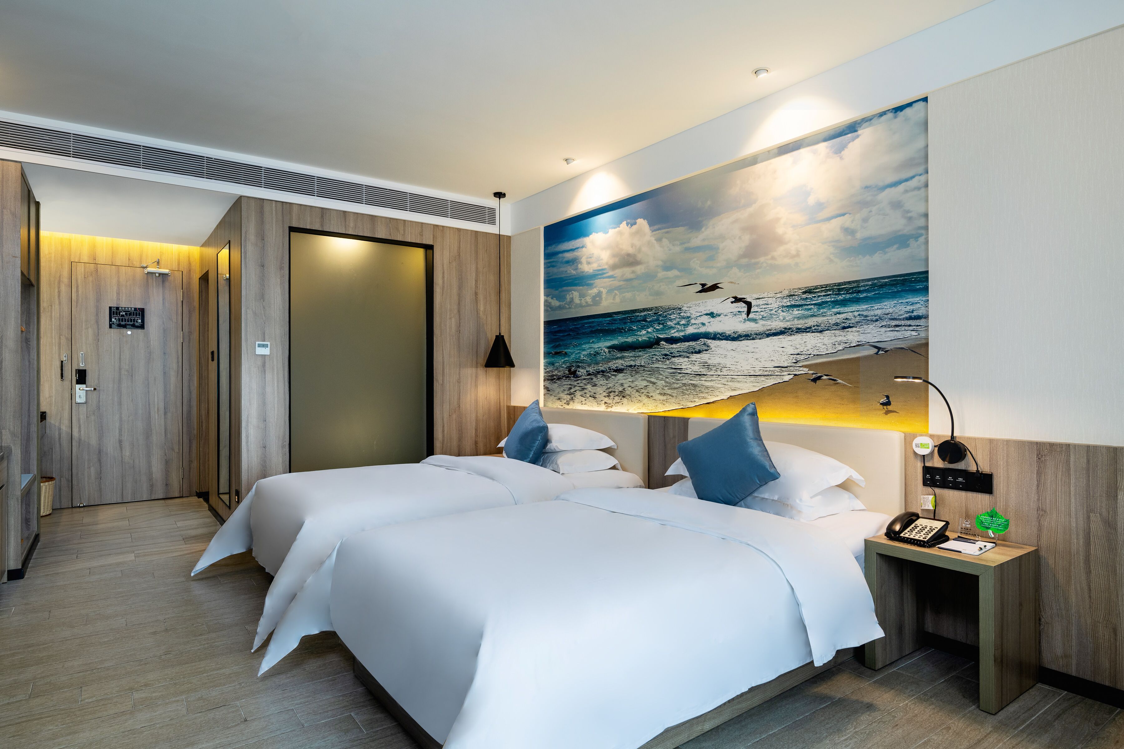 A hotel room with two beds, a painting of the ocean, a nightstand with a phone, and a wooden door.