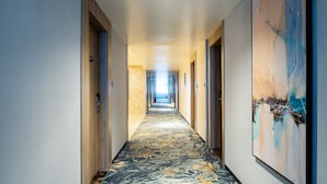 Un pasillo de hotel con alfombra estampada, puertas de madera y un cuadro abstracto grande en la pared derecha.