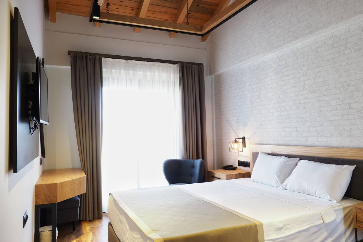 Quarto de hotel com cama, escrivaninha, cadeira, televisão e janela com cortinas.