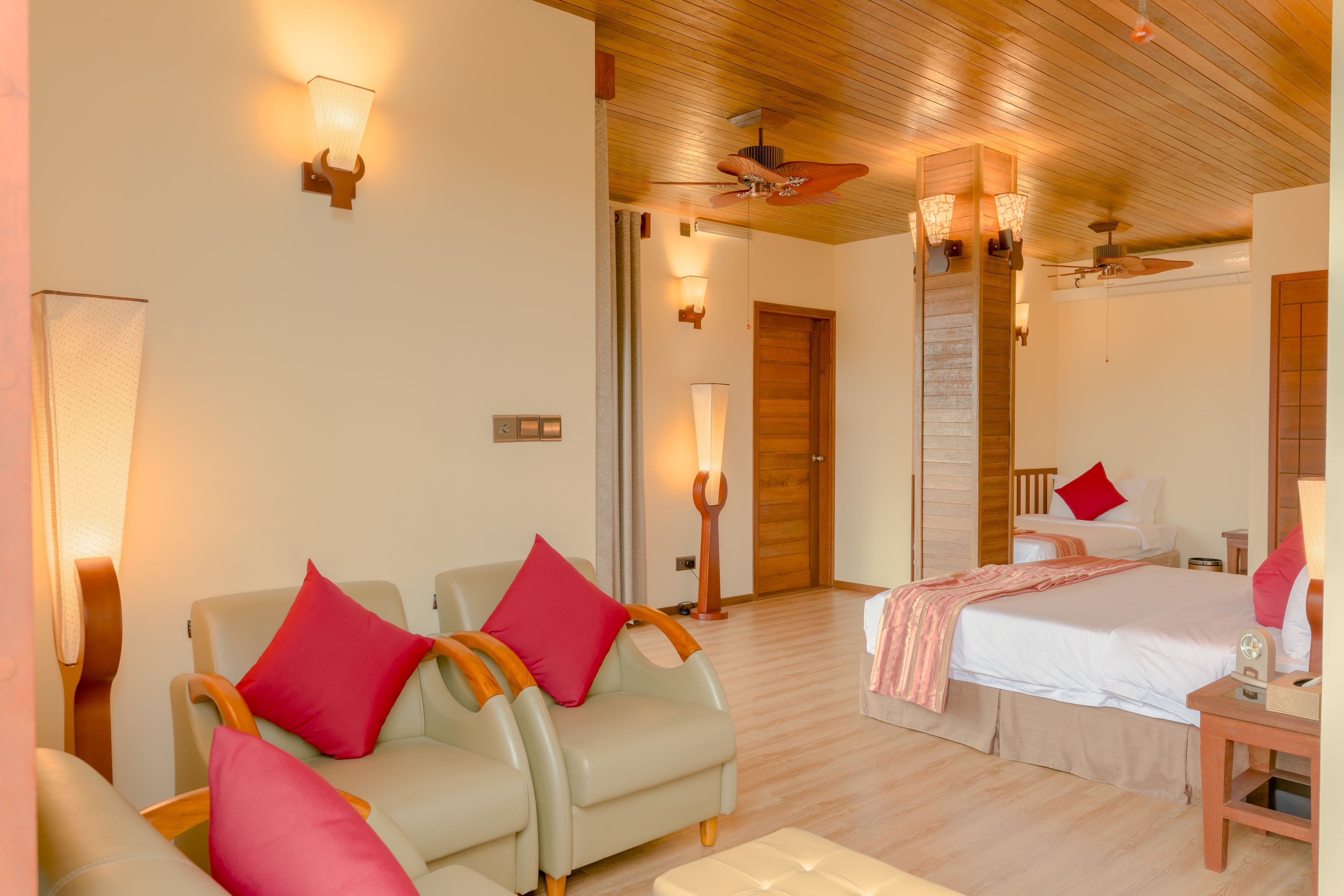A hotel room with two beds, a sitting area with chairs, a wooden ceiling, and wall-mounted lighting.