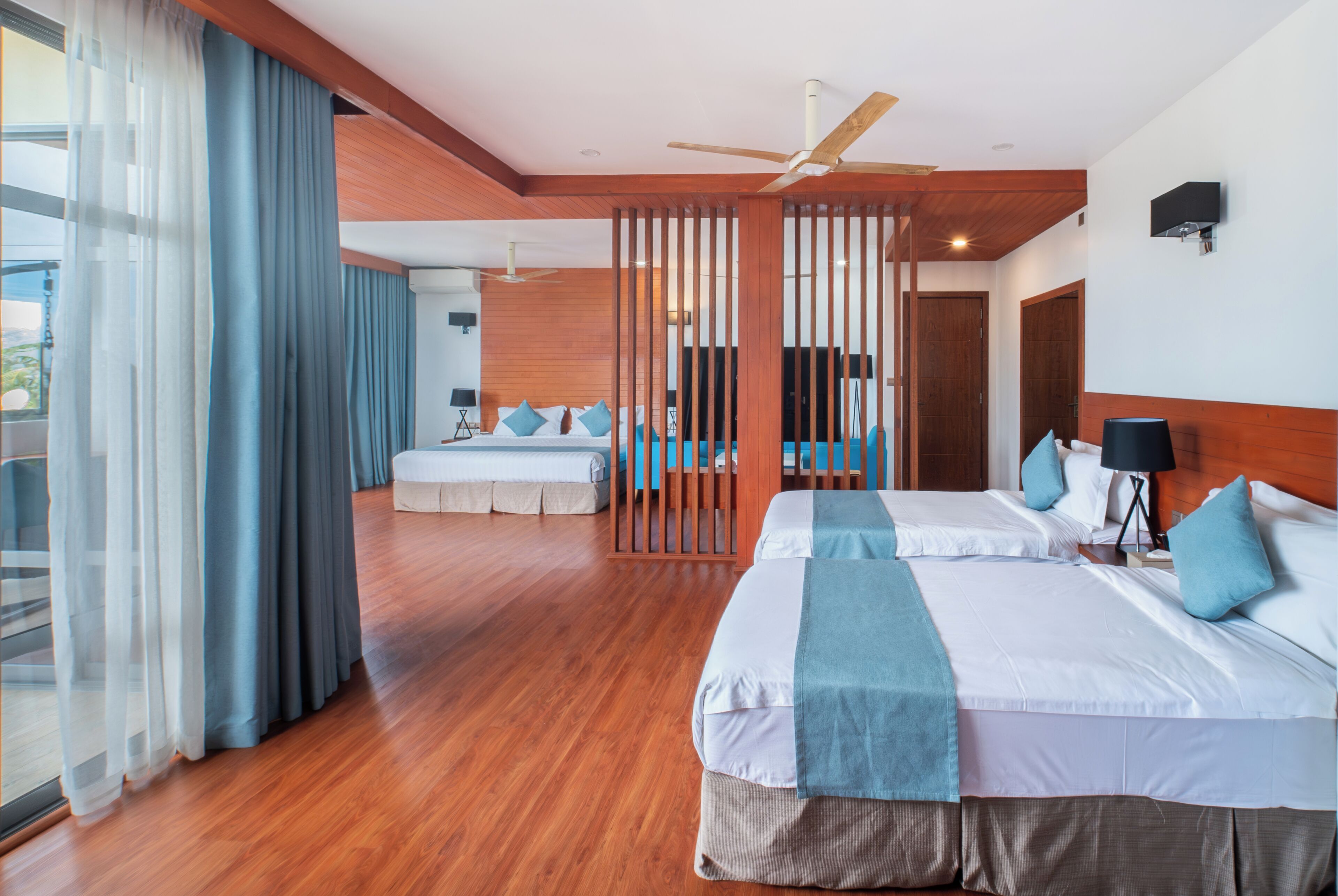 Habitación de hotel con dos camas, balcón, suelo de madera y ventilador de techo.