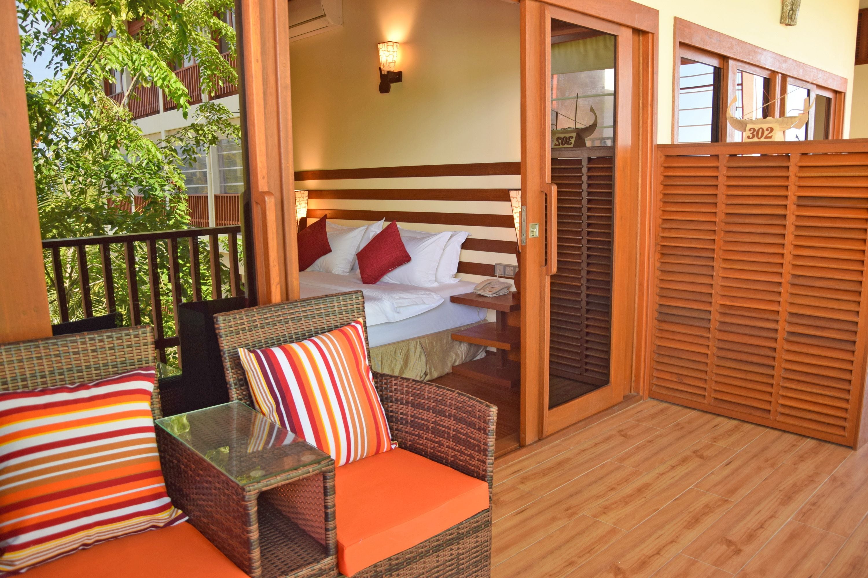 Un balcon agrémenté de meubles en osier, un lit avec des coussins rouges et blancs, et une porte en bois donnant sur une pièce.