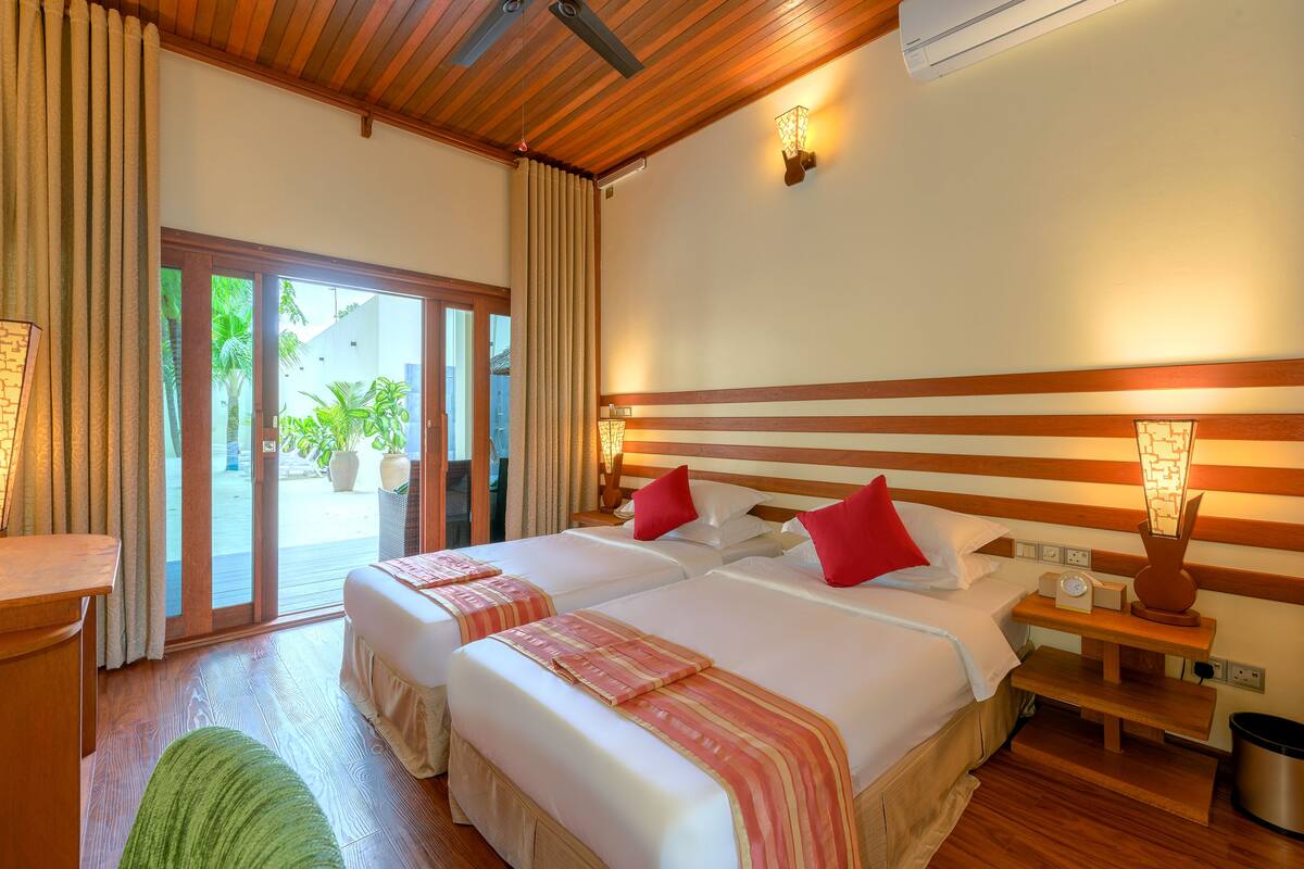 A hotel room with two beds, wooden flooring, a green rug, a wooden headboard, bedside tables with lamps, and a view of the outdoors through a sliding door.