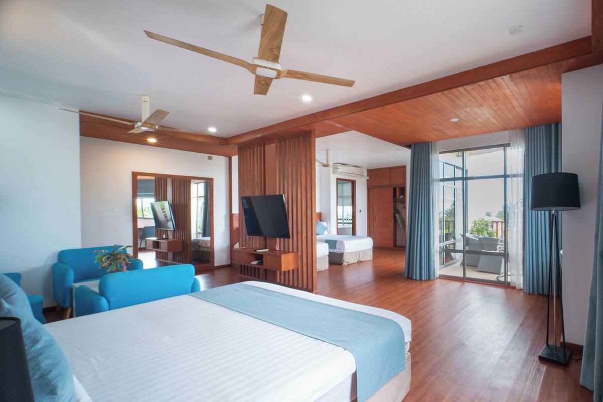 A spacious hotel room with a large bed, a seating area with a blue sofa, a flat-screen TV, wooden ceiling fans, and a balcony with a view.
