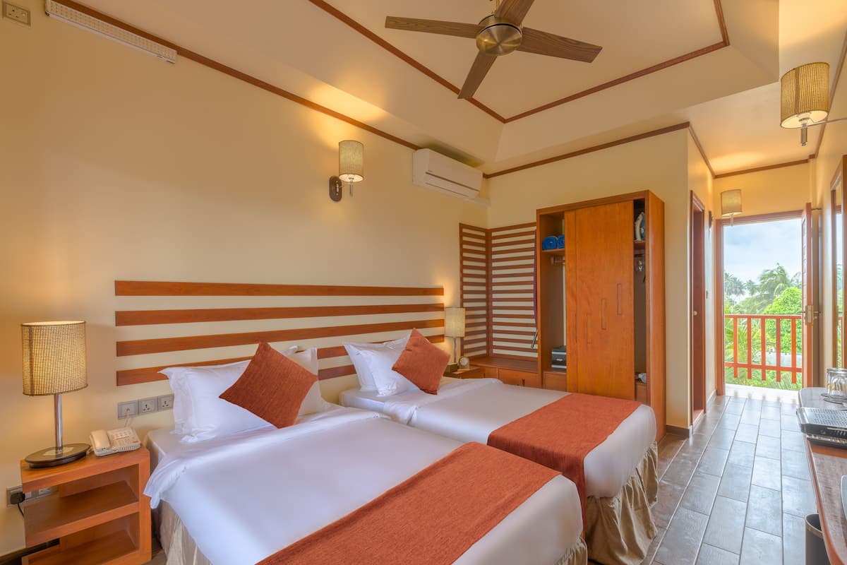 A hotel room with two beds, a ceiling fan, an air conditioning unit, a wooden wardrobe, and a balcony with a view of greenery.