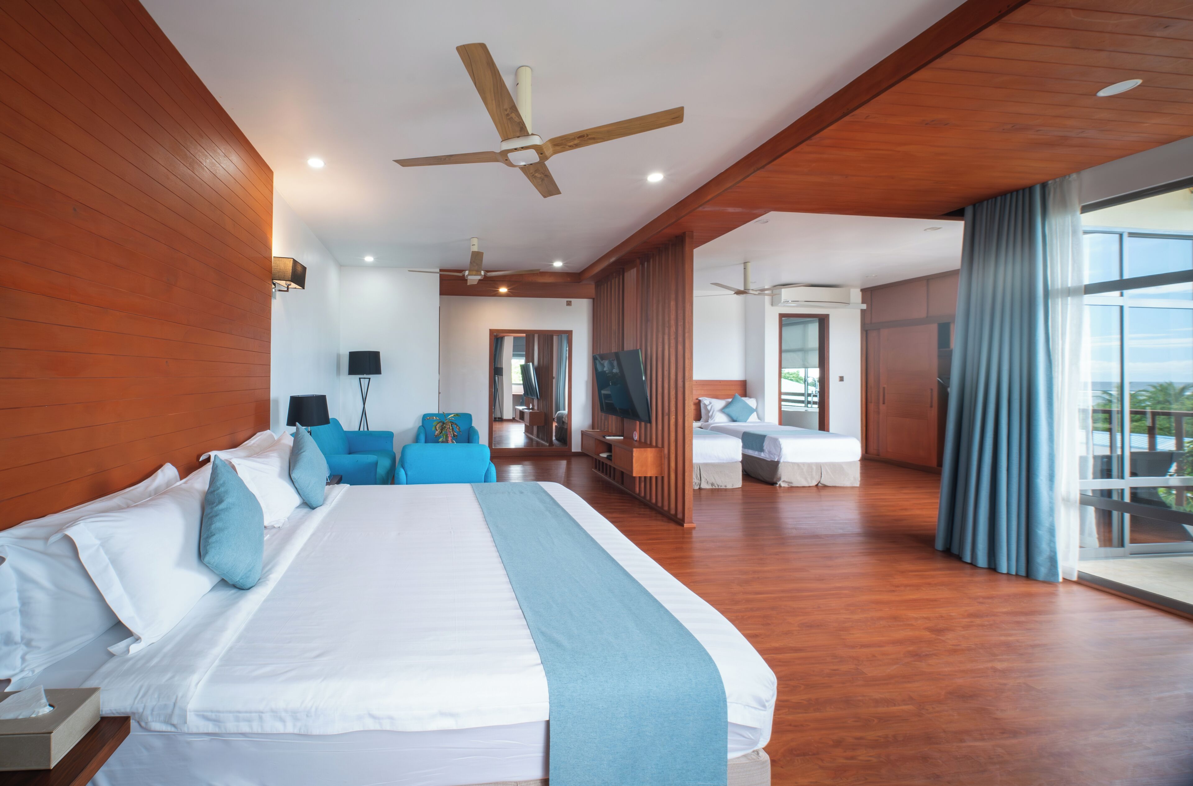 A spacious hotel room with a large bed, wooden walls, a ceiling fan, and a balcony with a view.